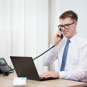 Junger Mann mit Brille telefoniert im Büro am Schreibtisch mit Laptop und Monitor.