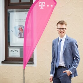 Junger Mann im Anzug steht vor Telekom-Shop mit pinker Fahne und Werbeplakaten.
